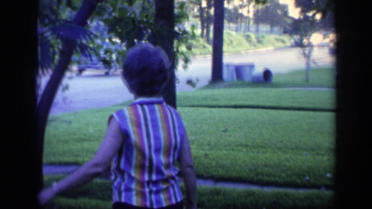 Woman seen from behind walking across a green front yard near trees and a street.