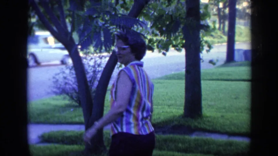A woman in glasses and a striped sleeveless blouse turns toward the camera in a leafy Florida suburban yard, with a street behind her.
