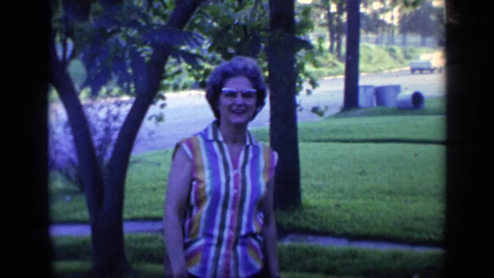 A smiling woman in eyeglasses faces the camera in a 1968 Florida suburban front yard, framed by trees, lawn, and a quiet road.