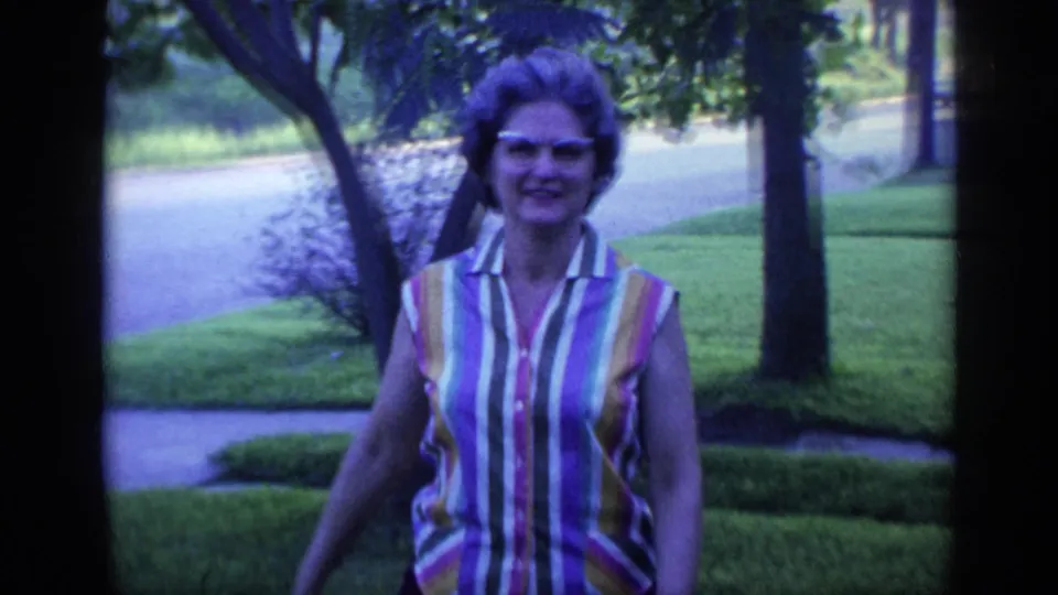 The woman smiles near the camera in a striped sleeveless top, with a Florida suburban lawn, trees, sidewalk, and road visible behind her.