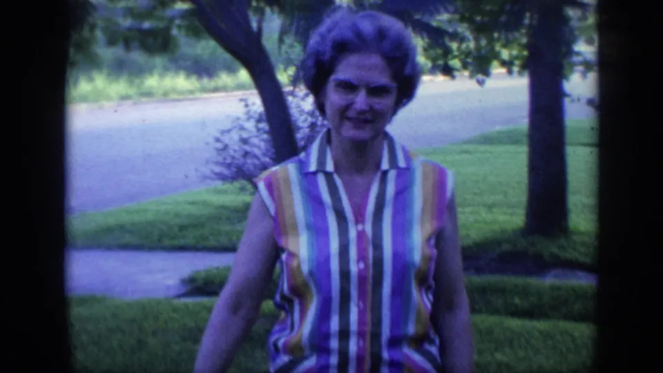 In 1968 Florida home-movie footage, a woman in a striped sleeveless shirt approaches the camera across a lawn near trees and a sidewalk.