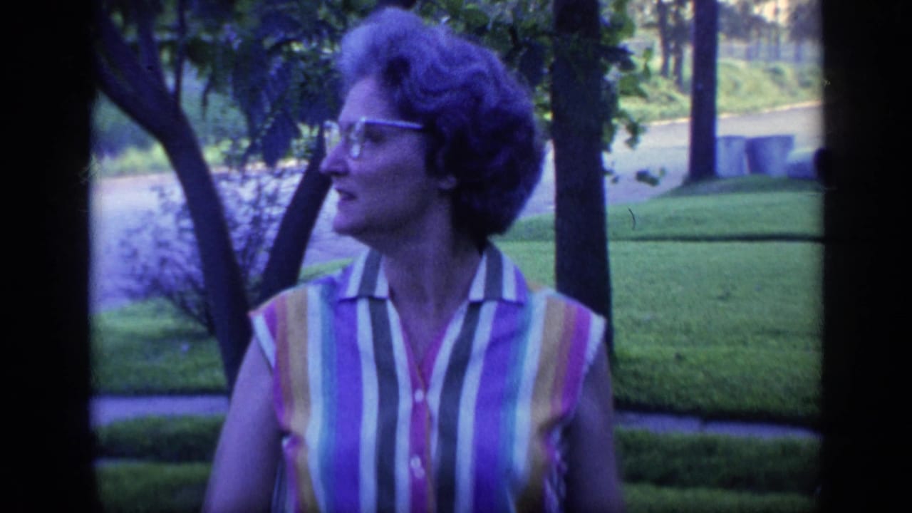 Woman in glasses turns in profile while standing on a lawn with trees and a road behind her.