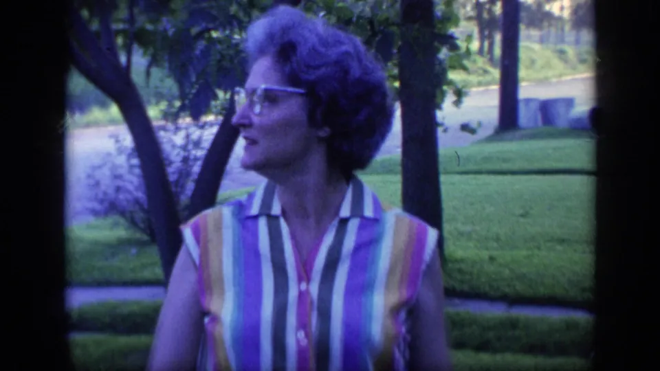 A woman wearing cat-eye glasses and a striped sleeveless blouse turns her head in a Florida suburban yard, with trees and a road behind her.
