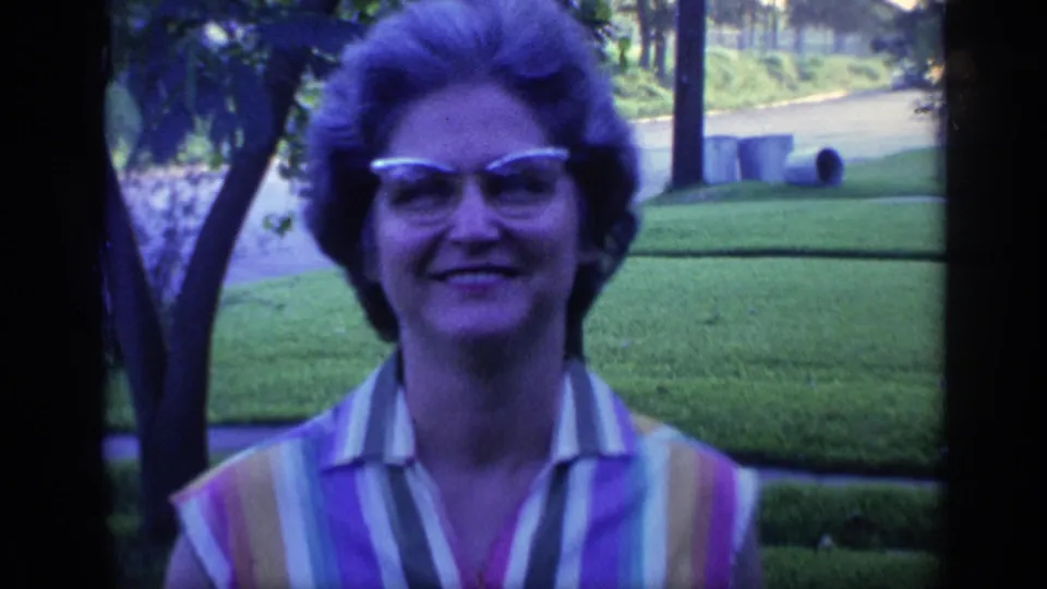 Close 1968 Florida home-movie view of a smiling woman in glasses and a striped sleeveless shirt, framed by lawn, trees, and roadside background.