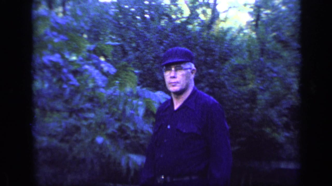 Man in a dark cap and shirt standing among dense green foliage.