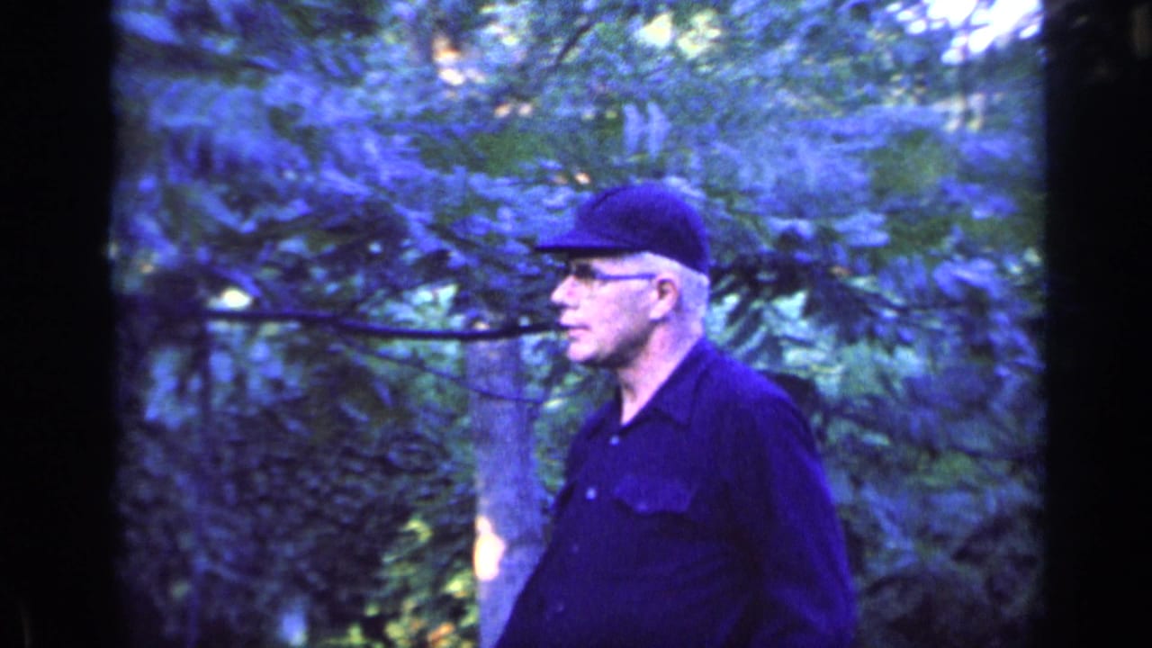 Man in profile wearing a dark cap while walking past trees and foliage.