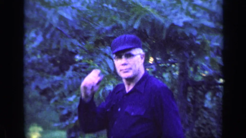 The man lifts a hand toward his cap, partly blurred by motion and the soft 8mm image.