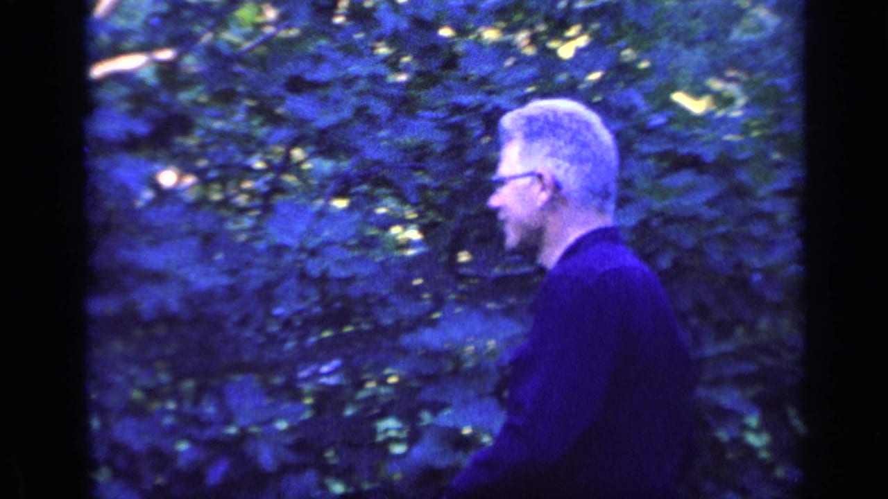 Gray-haired man in glasses stands in dense blue-cast foliage.