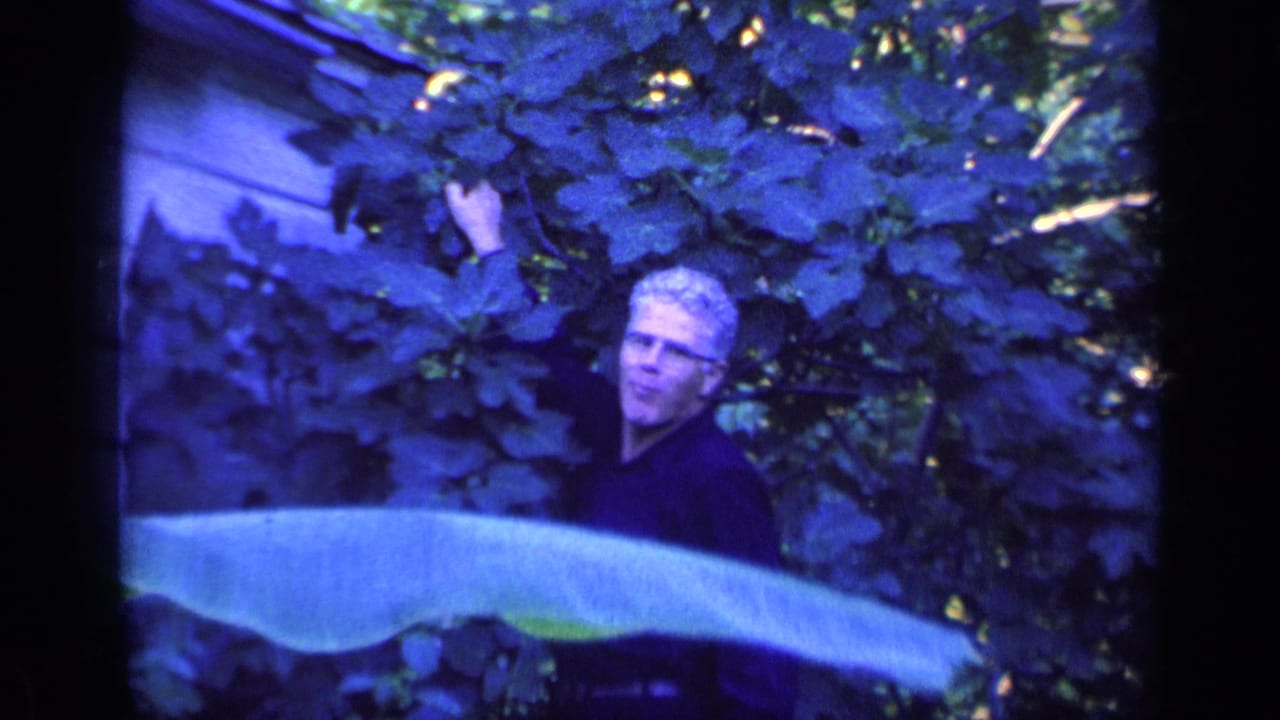 White-haired man partly hidden by broad leaves in a blue-tinted garden frame.