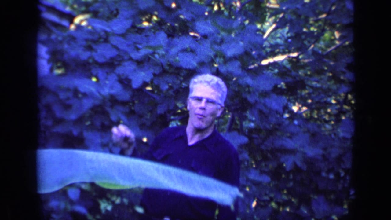 Man with glasses gestures near leafy branches in a garden.