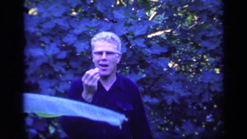 A white-haired man in glasses gestures with one hand toward the camera, with broad leaves and dense greenery around him.