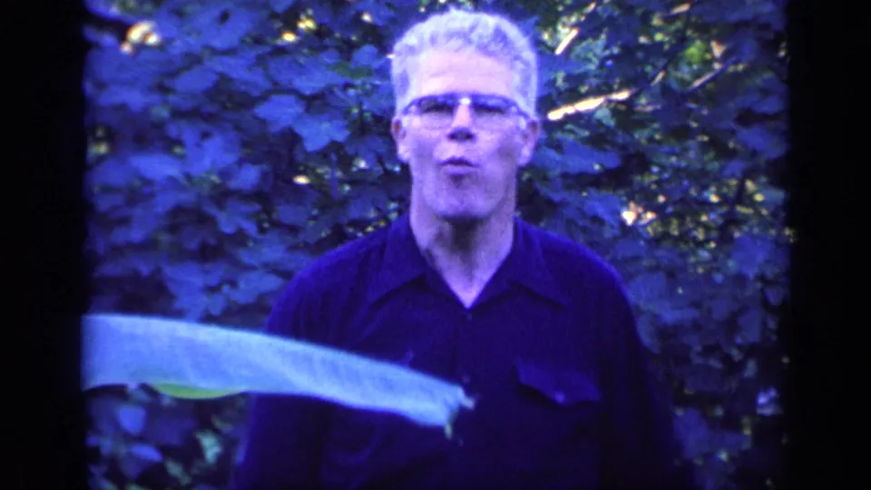 The man faces the camera in a closer 1968 Florida garden view, surrounded by foliage and a large leaf across the lower frame.