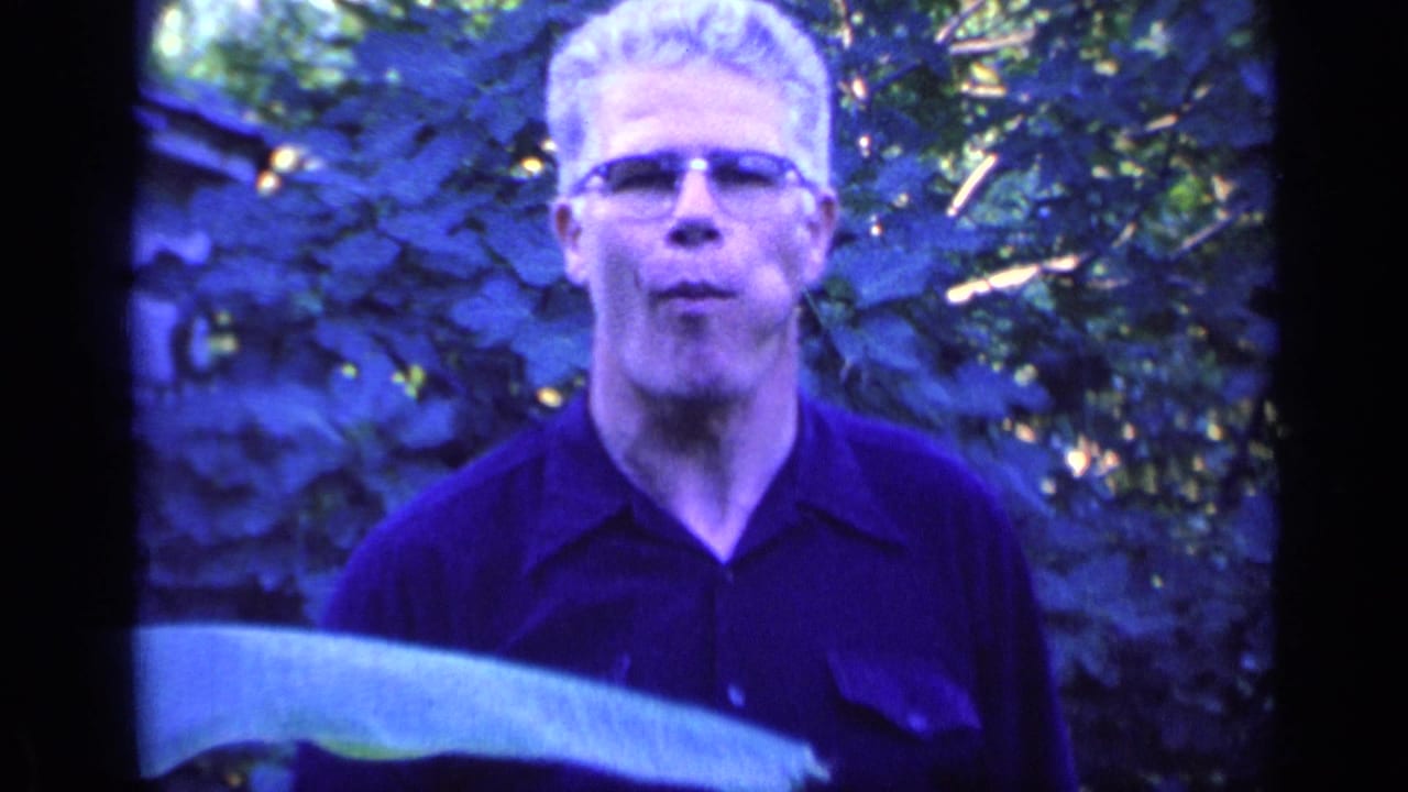 White-haired man with glasses stands outdoors in front of dense foliage.