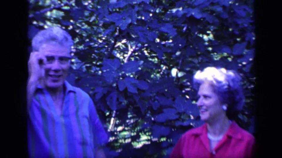 The man gestures near his glasses while the woman in red looks on smiling in the supported 1968 Florida home-movie setting.