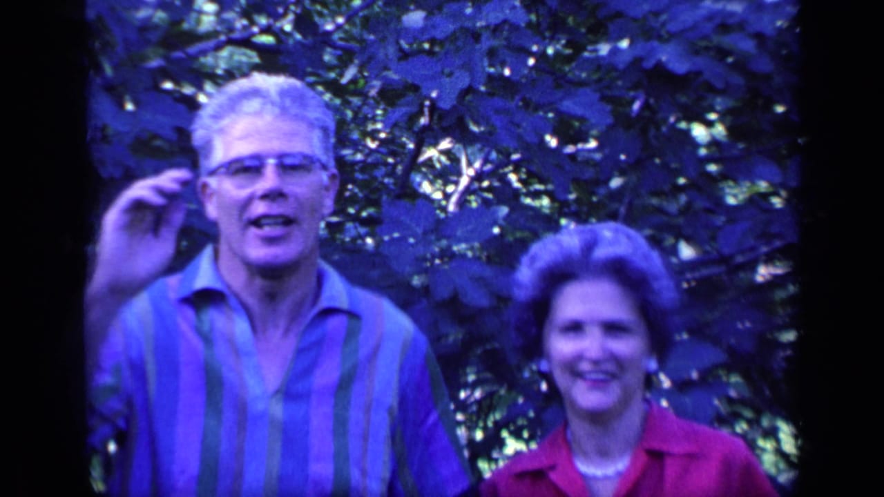 Older man with glasses gestures beside smiling older woman in leafy garden.