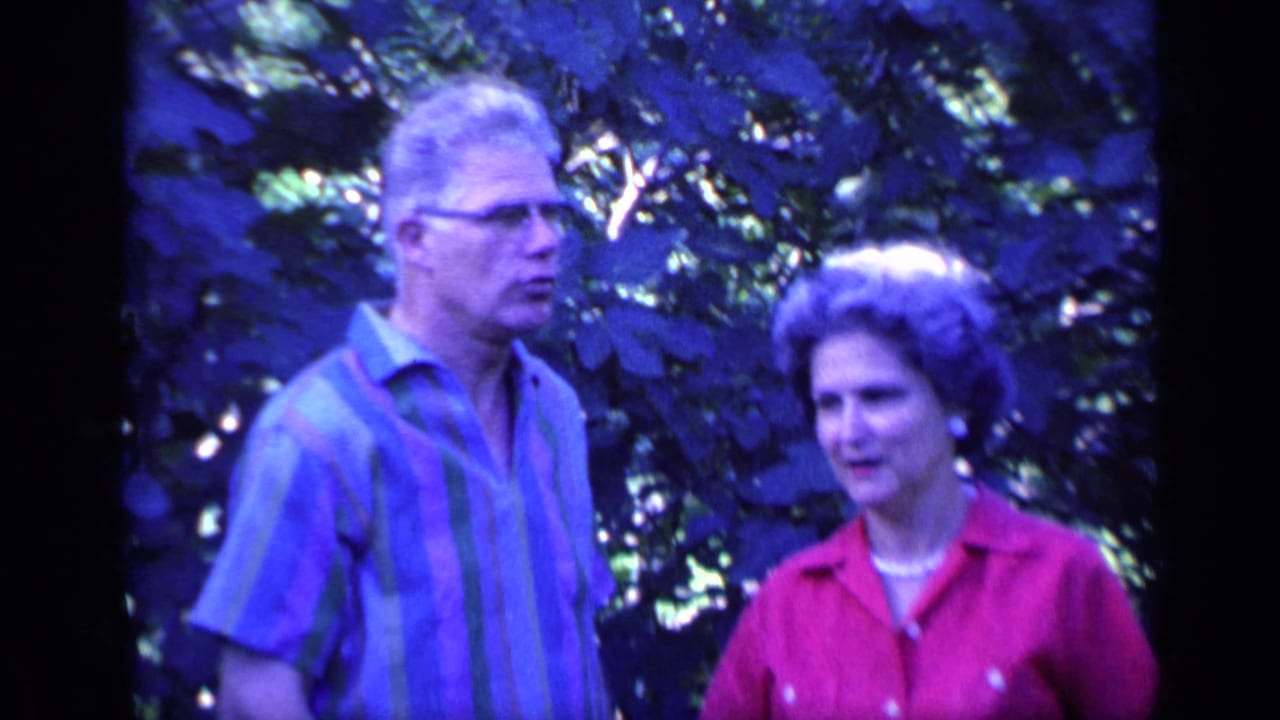 The older couple stands in a leafy Florida garden setting, with the man turned slightly away and the woman facing toward the camera.
