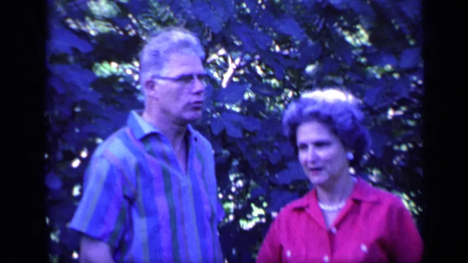 The older couple stands in a leafy Florida garden setting, with the man turned slightly away and the woman facing toward the camera.