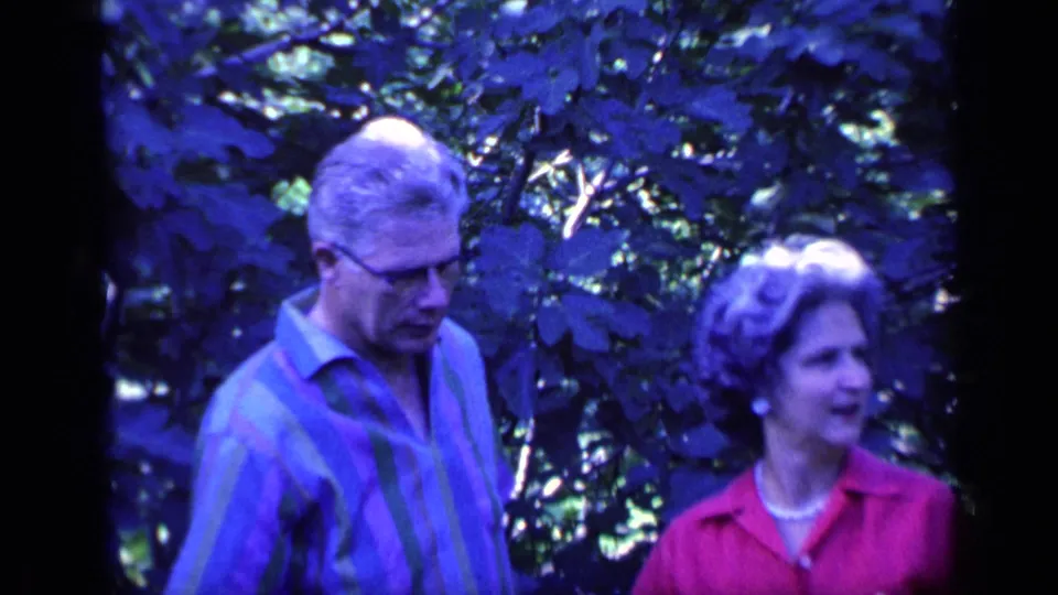 In the 1968 home-movie frame, the man lowers his gaze as the woman stands beside him against dense garden leaves.
