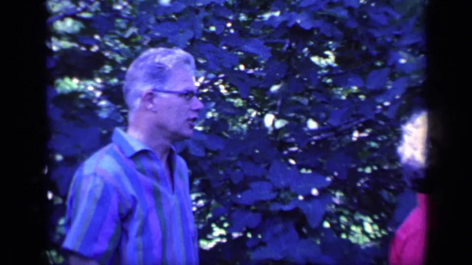 The man in glasses turns to the right in the leafy garden, while the woman in red is only partly visible at the frame edge.