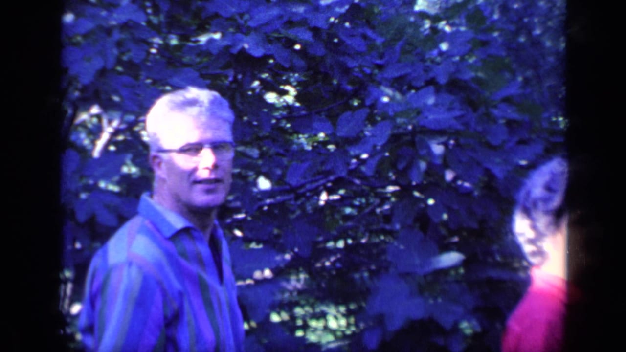 Man with glasses in a striped shirt stands outdoors near leafy trees.