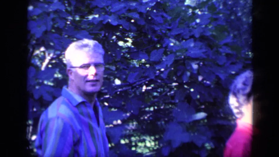 In 1968 Florida home-movie footage, a man with glasses stands in a shaded garden beside dense foliage.