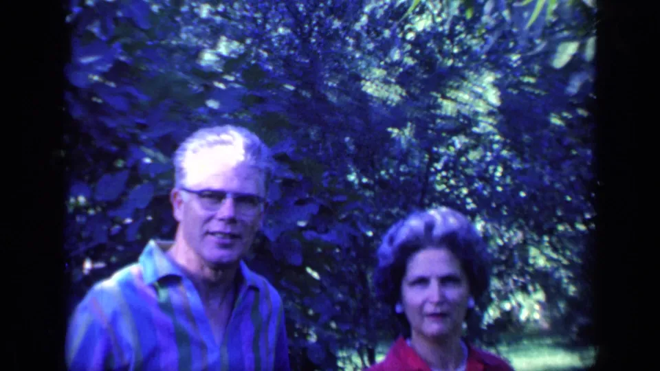 A middle-aged couple faces the camera in 1968 Florida footage, framed by shaded trees and bright lawn beyond.
