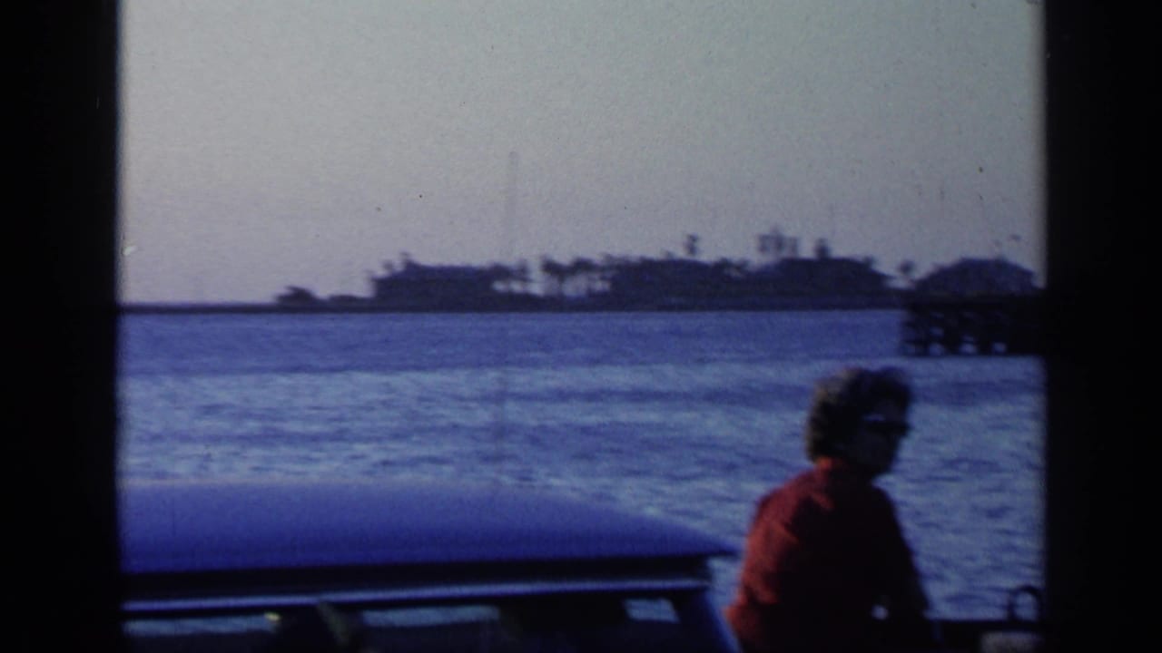 Woman in a red shirt sits on a boat with shoreline across the water.