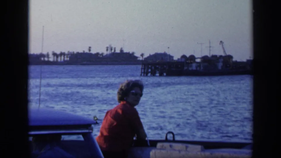 1968 Florida boat passenger in sunglasses sits near the rail as the camera looks toward water, pier structures, and palm-lined shore.