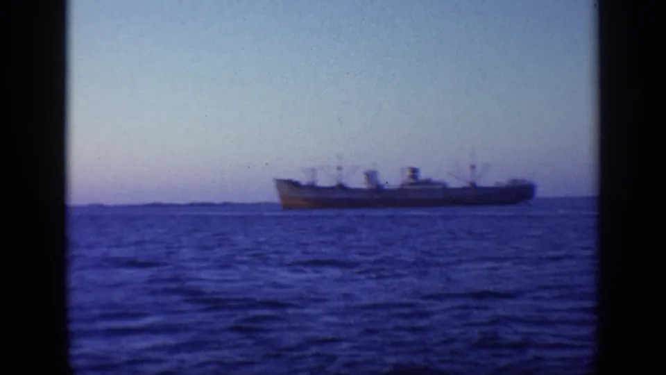 1968 Florida 8mm frame showing a large cargo ship in profile on calm blue water, with a low shoreline and fading dusk light.