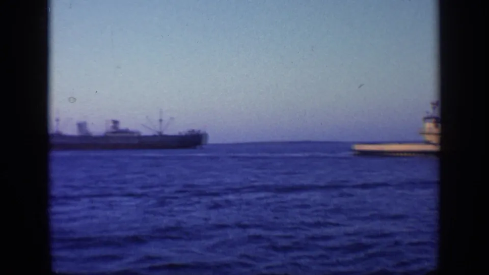 1968 Florida 8mm frame showing a cargo ship offshore, low land on the horizon, and a small pier or vessel edge entering from the right.