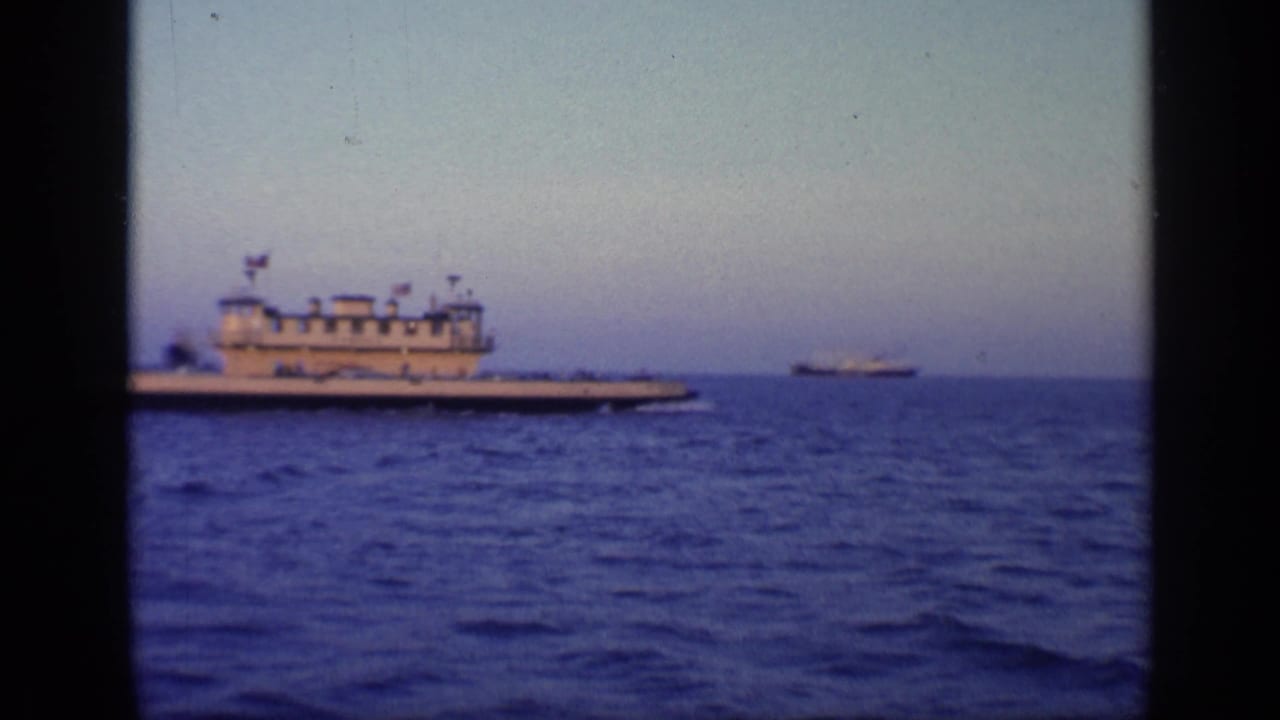White multi-deck vessel crossing blue ocean with pale sky and film borders.