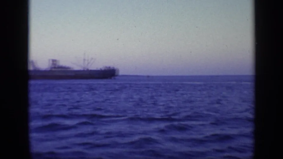1968 8mm frame from Florida metadata showing a distant cargo ship crossing blue open water beneath a pale horizon.