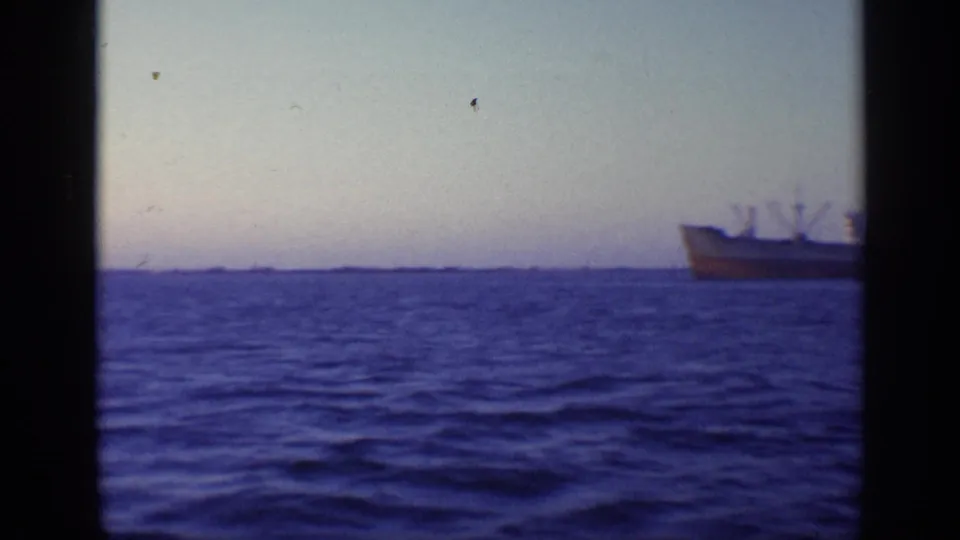The camera holds on blue water and a low horizon while the cargo ship slips partly out of frame at the right edge.