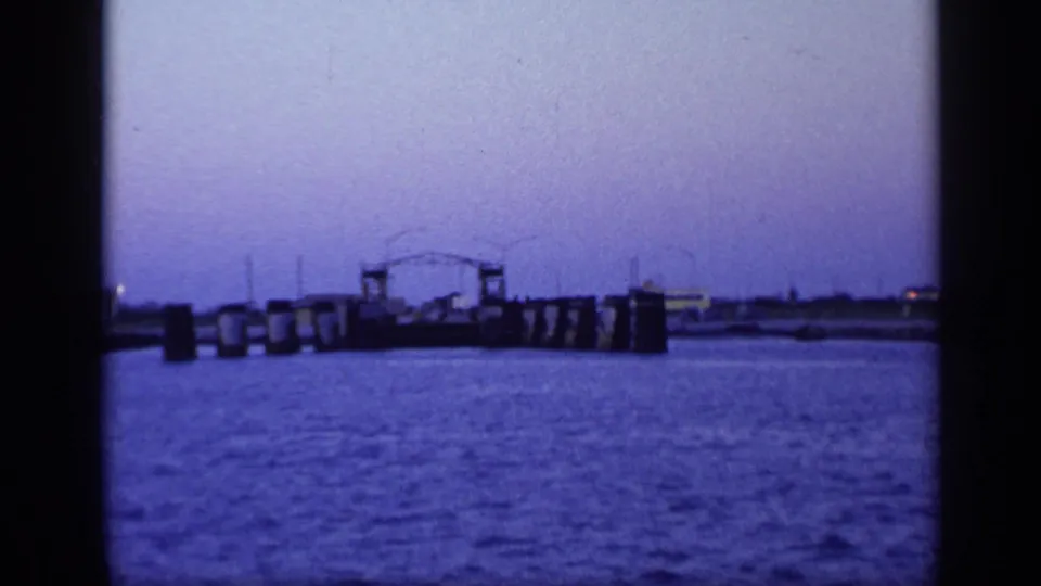 1968 Florida 8mm frame showing harbor water, pier pilings, and a distant low industrial or marina structure.