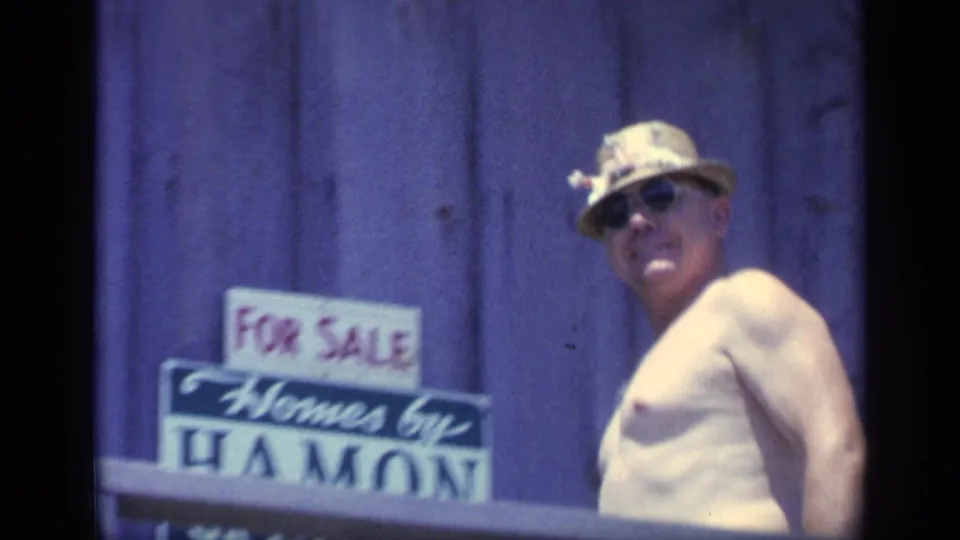 1968 Florida home-movie frame of a shirtless adult man in a hat and sunglasses near a wooden building with sale signage.