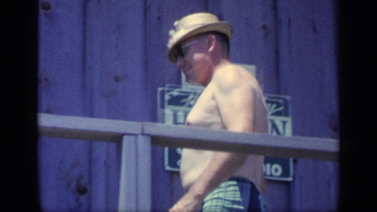 Shirtless man in a straw hat walking beside a porch railing with a sign behind him.