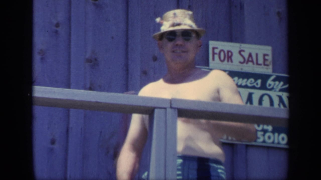 Man in sunglasses and straw hat stands near a porch railing with a For Sale sign behind him.