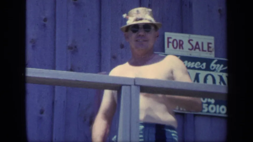 A shirtless man in sunglasses faces the camera on a Florida porch, with a For Sale sign partly visible on the wooden wall behind him.