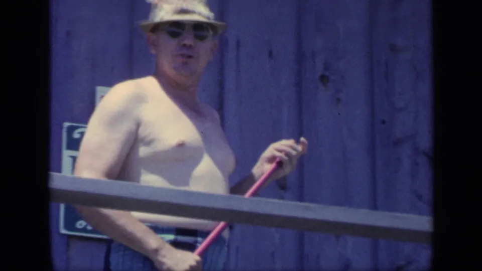The man looks toward the camera while holding a broom handle on the porch, framed by railing, purple siding, and a partially visible sign.