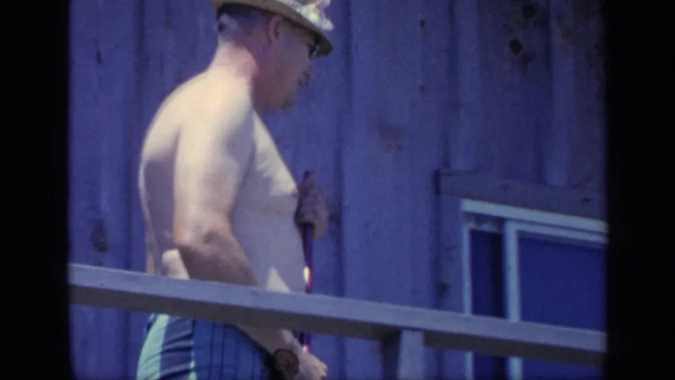 A side view shows the shirtless man holding the broom near the porch railing, with wooden siding and a small window visible in the frame.