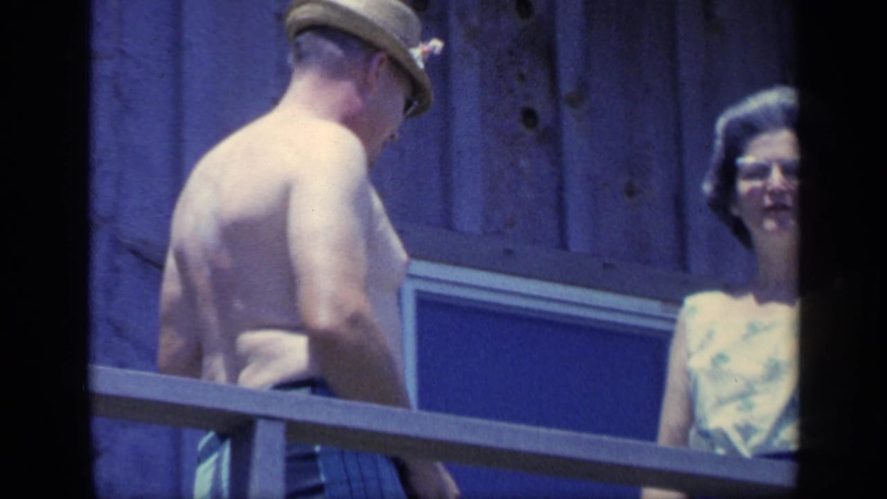 Shirtless man in a tan hat stands near two women behind a wooden balcony railing.
