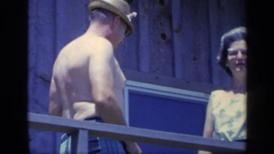 1968 Florida 8mm frame showing a shirtless man in a tan hat beside two women on a sunlit wooden balcony.