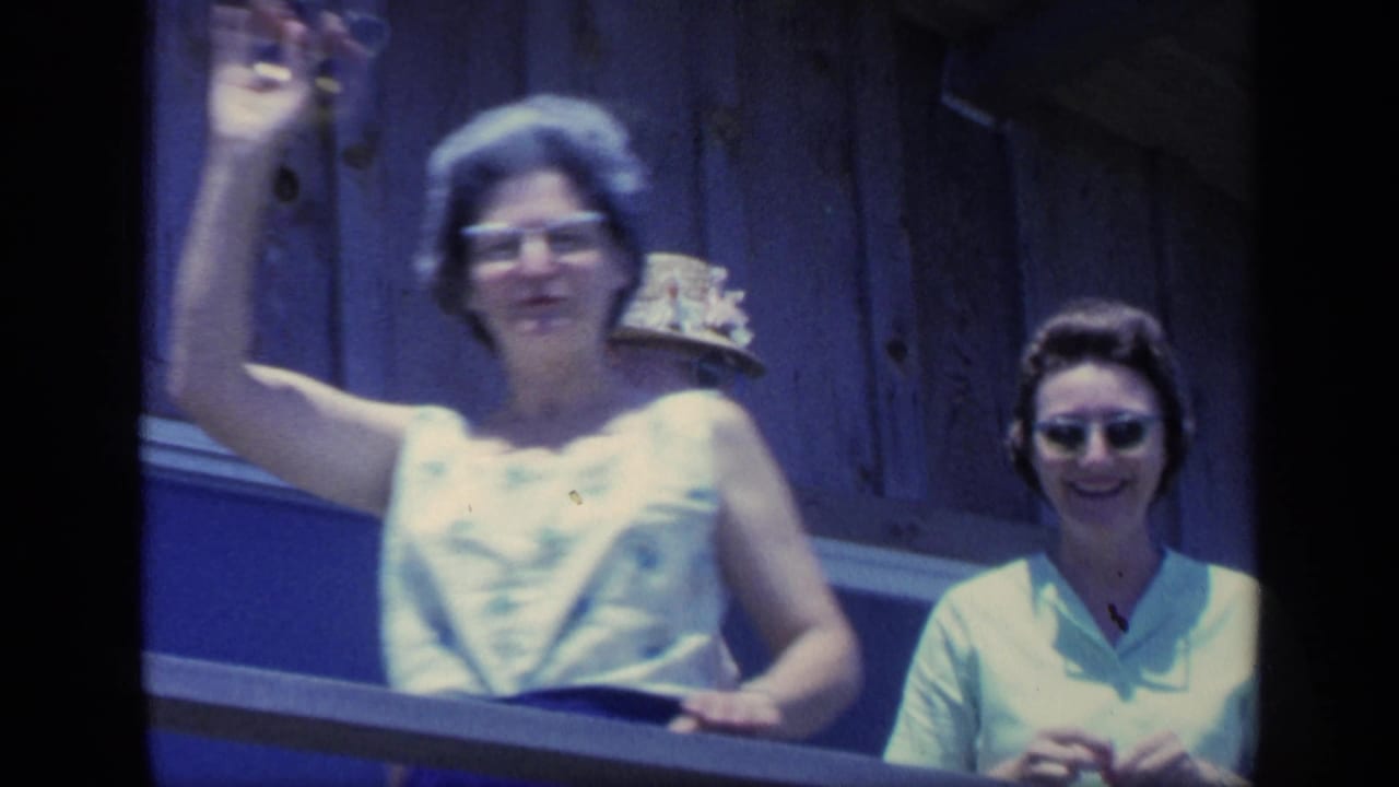 Woman in glasses waves from a wooden balcony while another woman stands nearby.