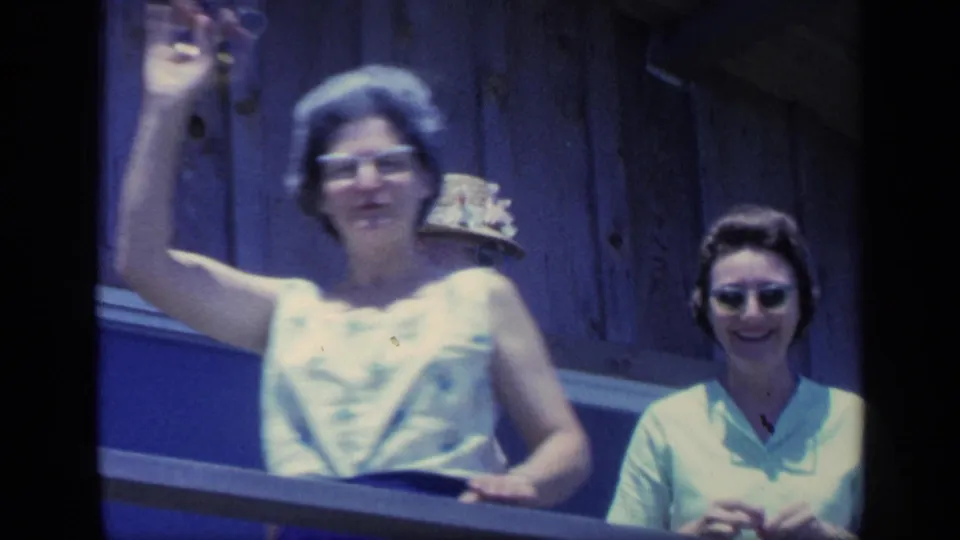1968 Florida home-movie frame of a woman in glasses waving from a balcony as another woman looks toward the camera.