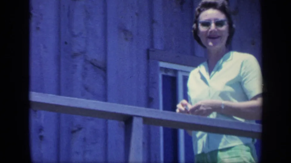 1968 Florida home-movie frame of a smiling woman in glasses on a sunlit wooden balcony.