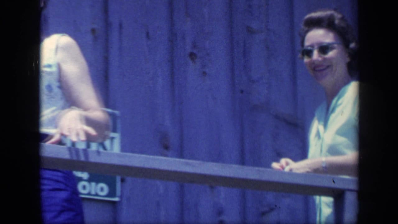 In 1968 Florida home-movie footage, a woman in sunglasses stands by a porch railing against blue wooden siding, with another person partly cropped at left.
