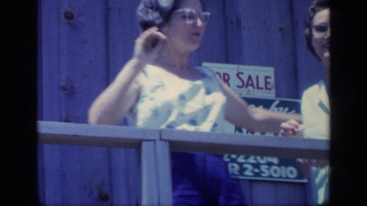 A woman in glasses and a sleeveless floral top gestures near a porch railing, with a partial For Sale sign behind her in the 1968 Florida clip.