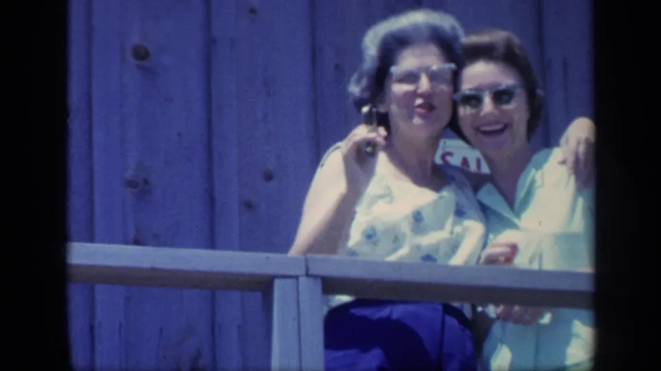 Two women in 1960s attire stand close together on a Florida porch, smiling toward the camera with blue siding and a partial sign behind them.