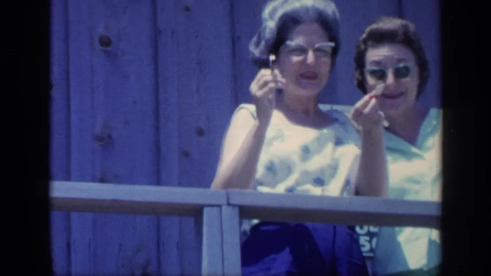 Two women on a Florida porch point and gesture toward the camera, one in glasses and a floral sleeveless top and the other in sunglasses.