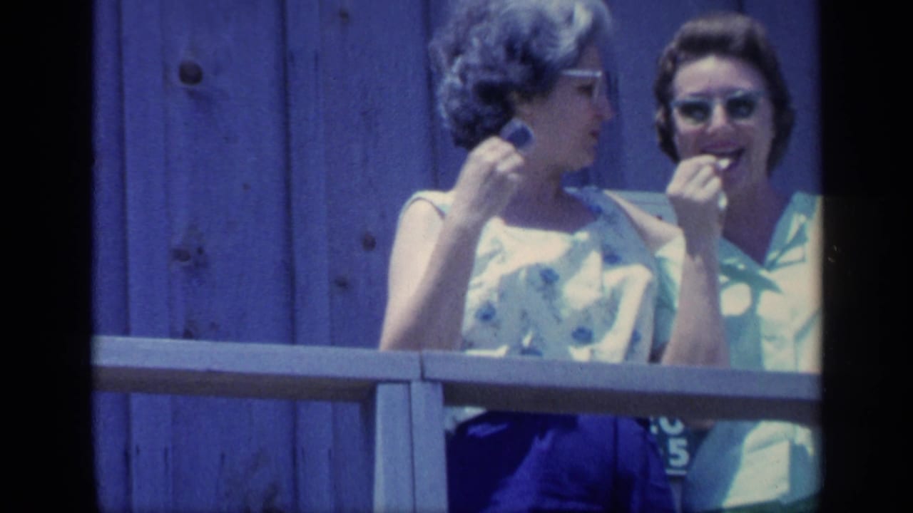 Two women stand behind a wooden railing, one wearing glasses and holding a snack.